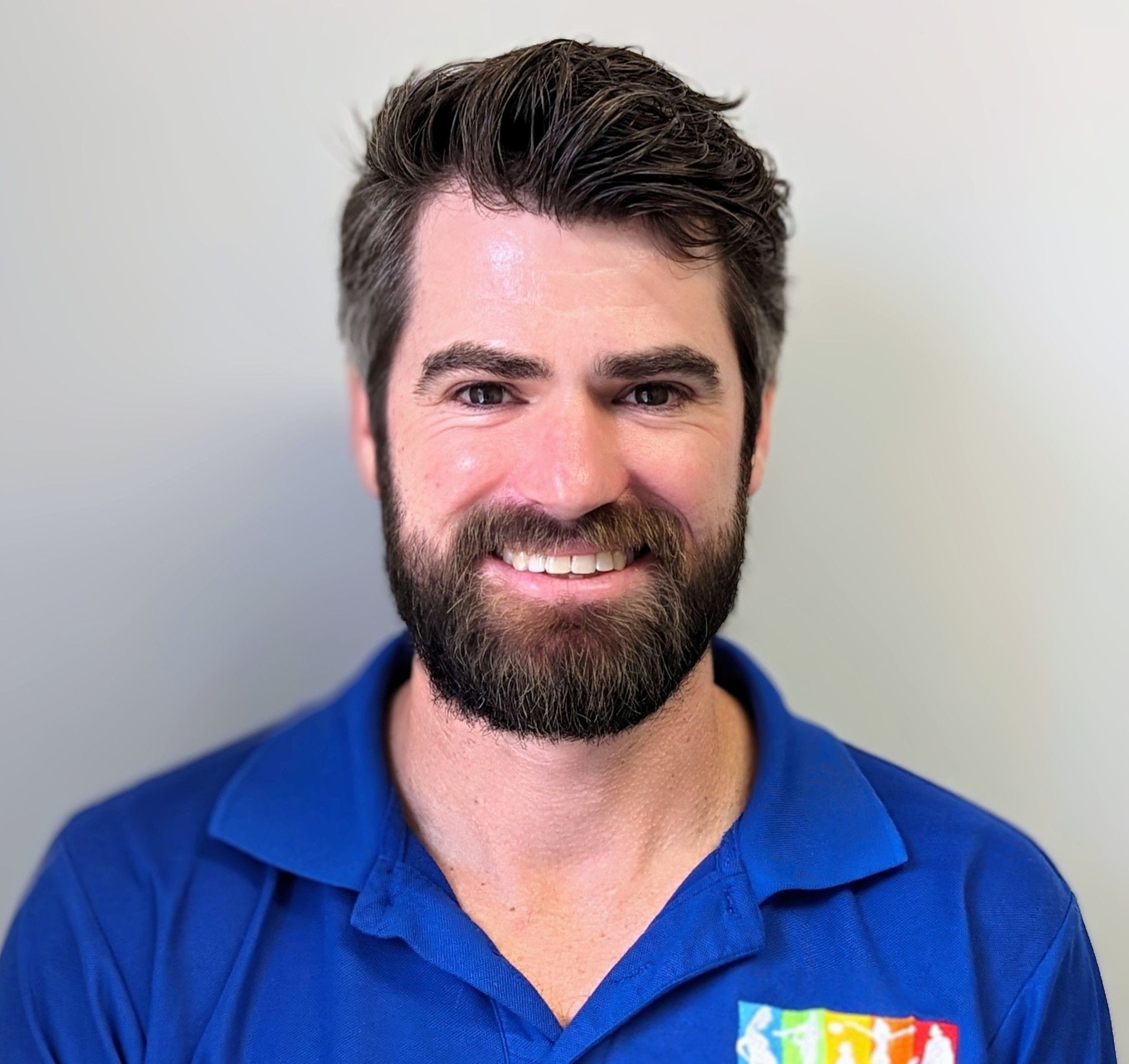 Staff photo of Logan Dolbel Thomas, wearing a checked blue and white shirt in front of a plain grey background.