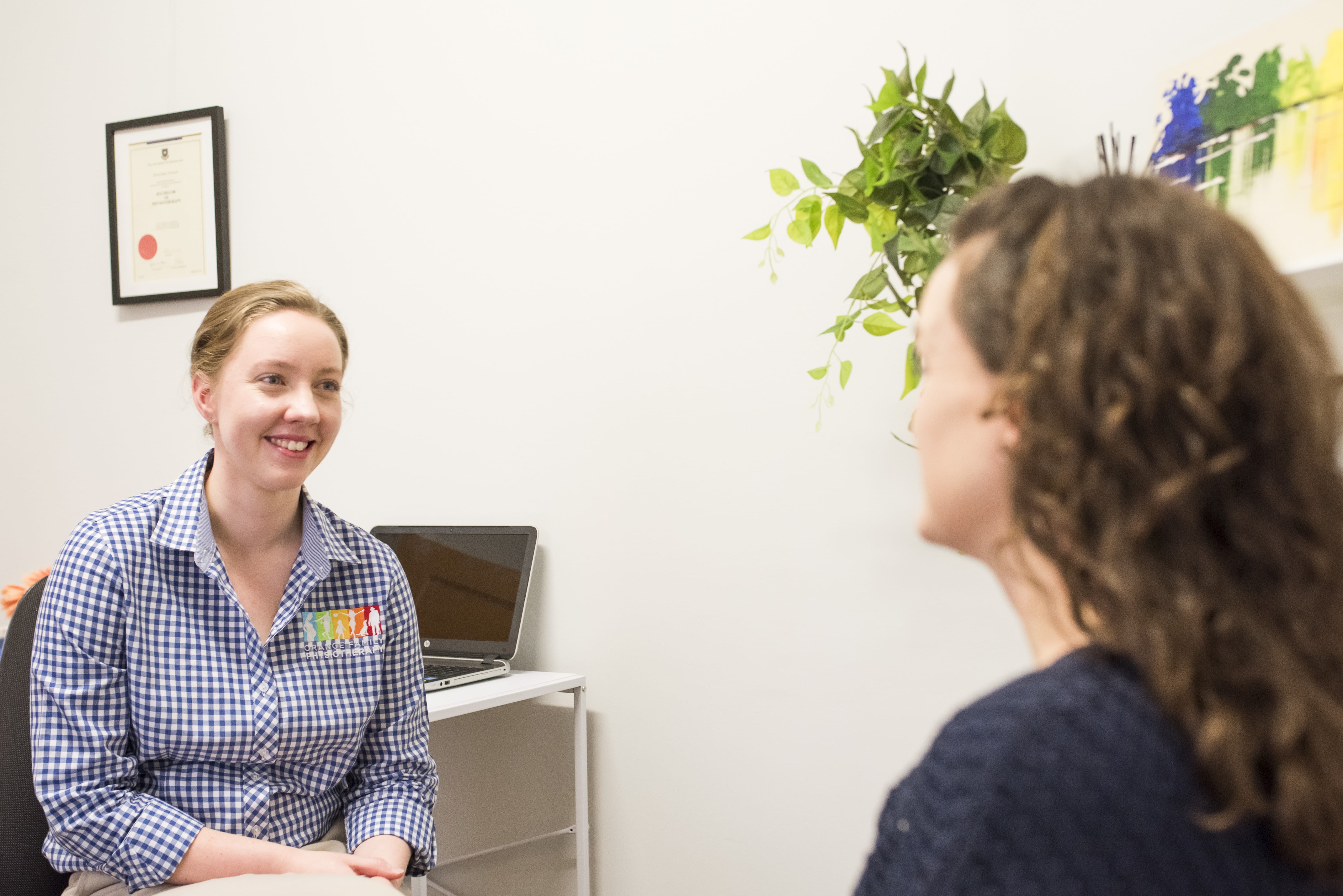 Physiotherapist Monica Carmen wearing checked blue shirt with Orange Family Physiotherapy logo speaks to a client.